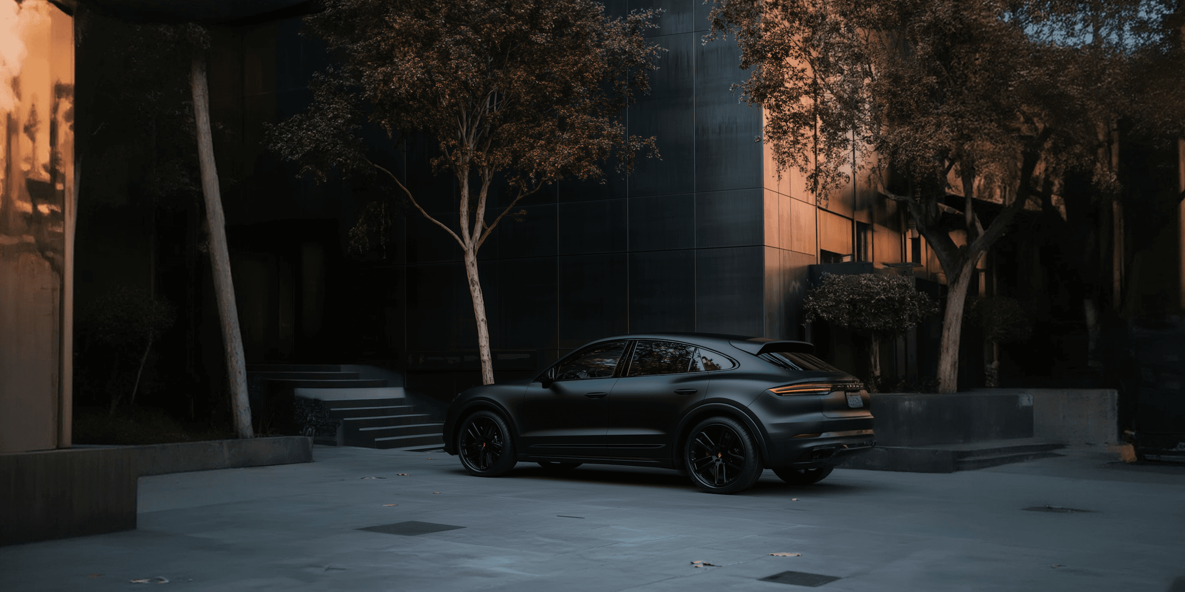 Matte black Porsche Cayenne Coupe parked in a modern urban courtyard with golden hour light.